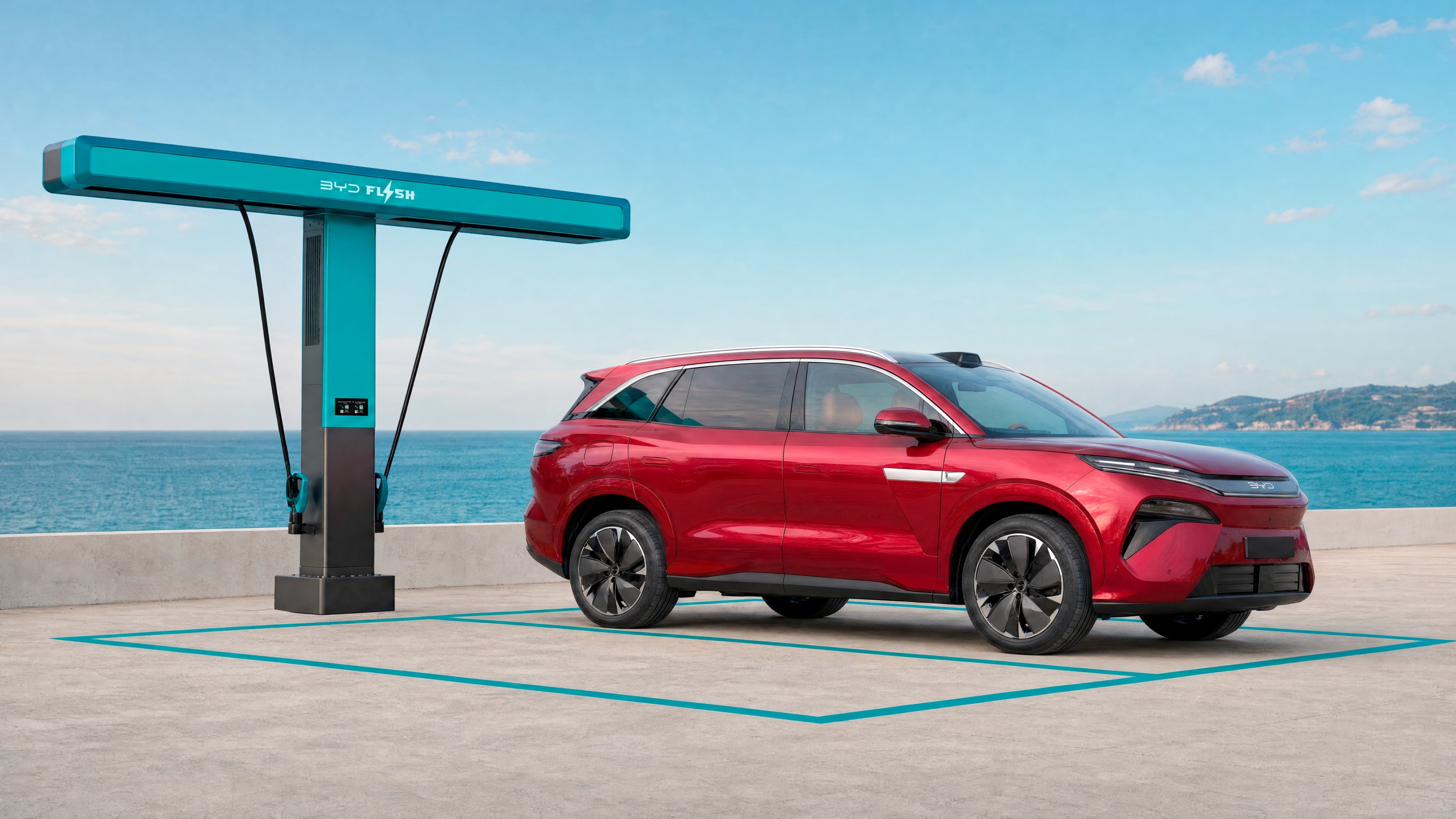 Red BYD Atto 8 in a parking space by the sea, next to a turquoise, T-shaped flash charger with two charging cables hanging down from the top.