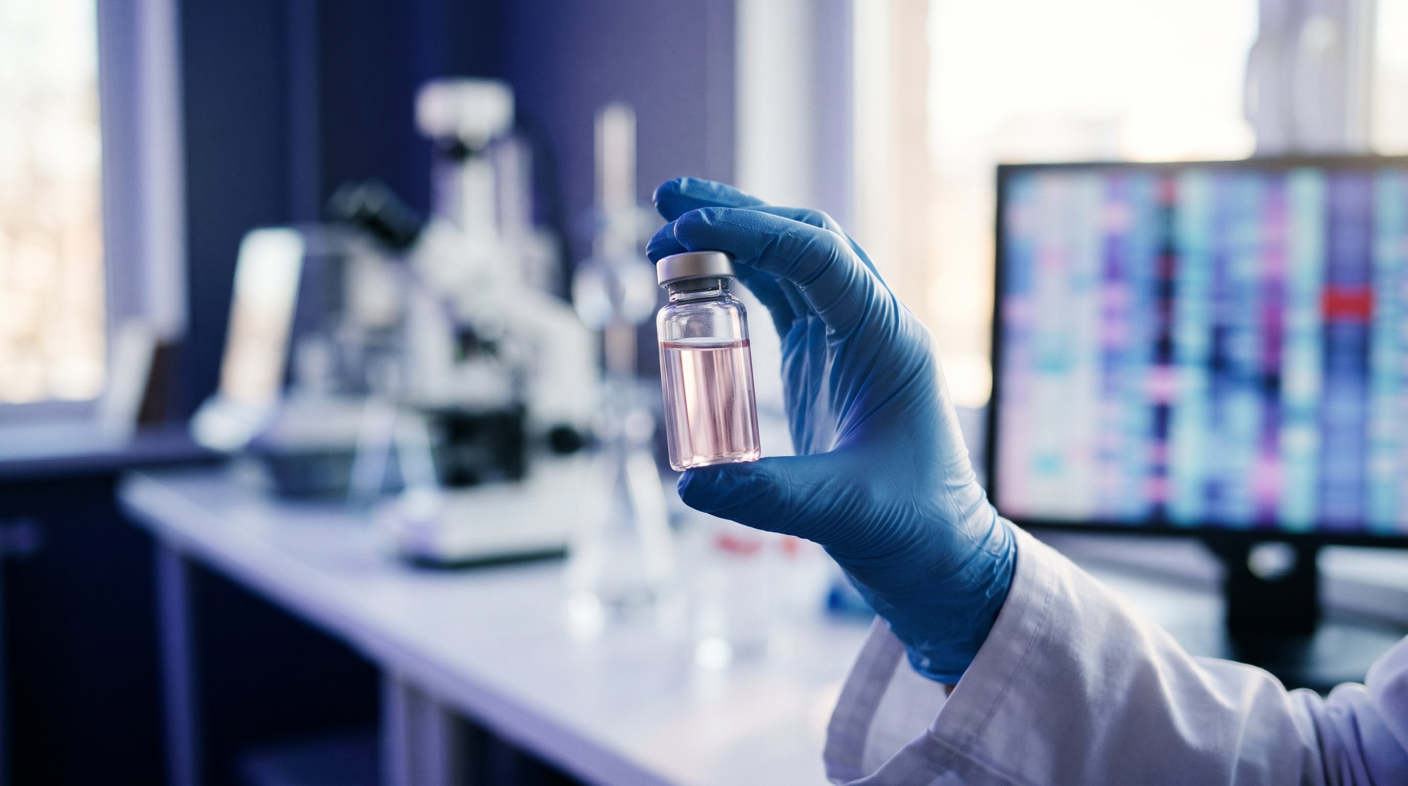A hand in a blue glove holds up a small glass vial with pale pink liquid in a lab, with a blurred screen full of coloured bands from a genetic analysis in the background.