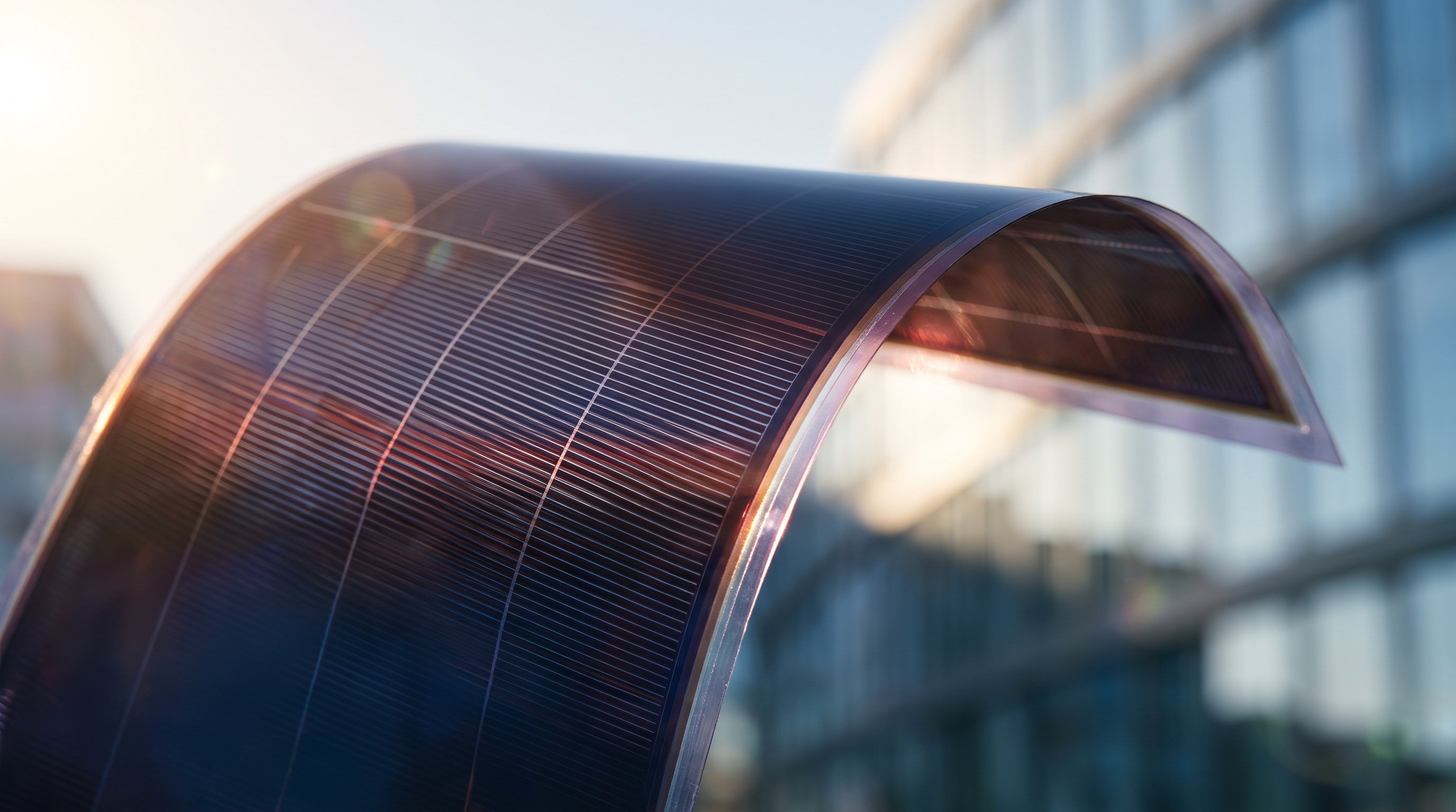 A thin, flexible solar panel with a fine grid of narrow cells, bent into an arch against the light of the sun, in front of a blurred glass facade.